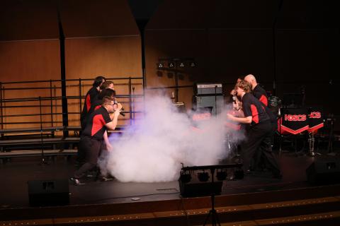 A group of singers on stage facing each other with smoke from a smoke machine bellowing between them.