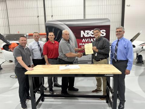 Group of 6 men standing around a table in an airport hangar. Two of the men are shaking hands and holding a certificate.